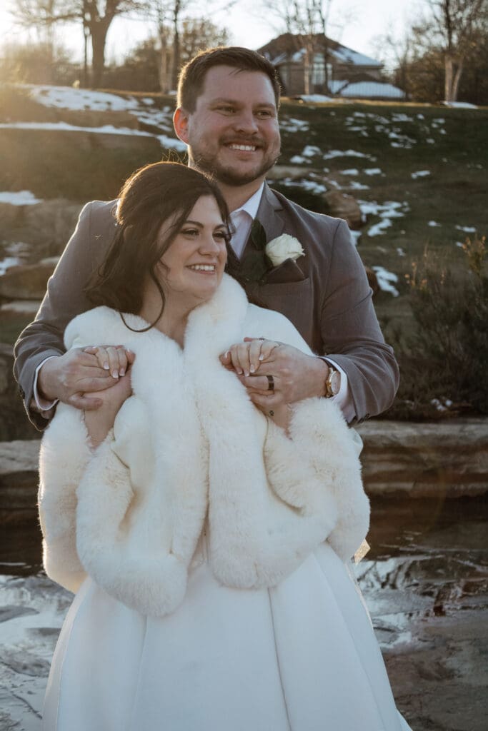 Groom holding brides hands from behind with snow and grass in the background